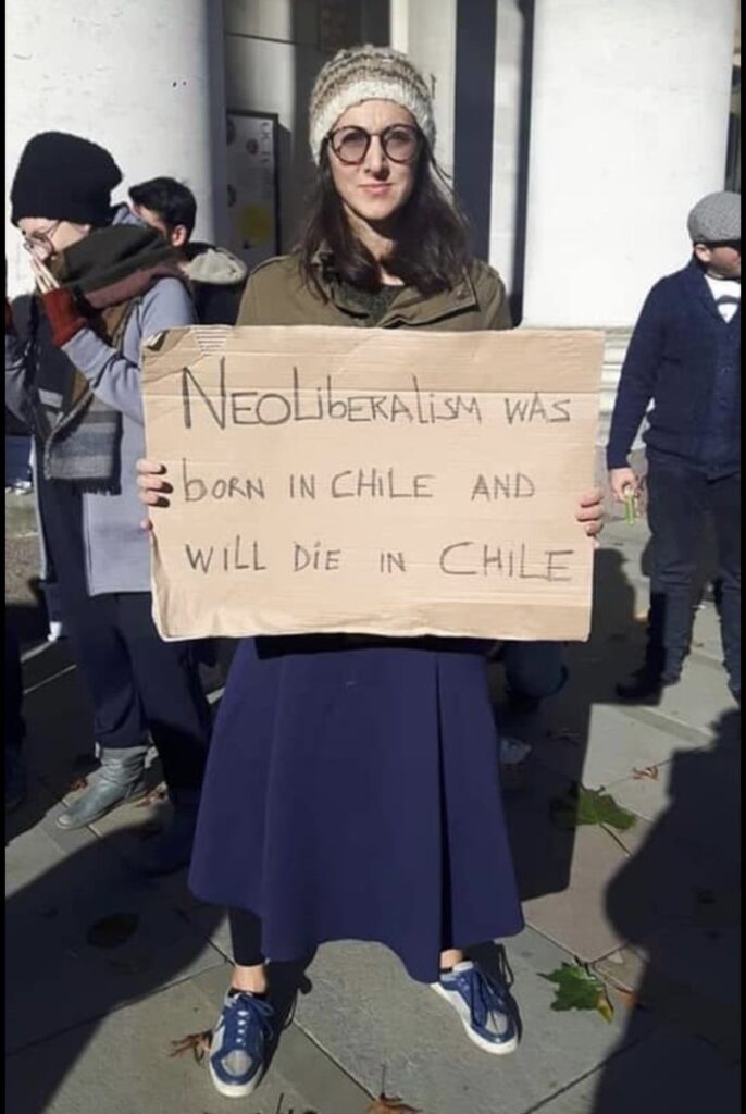 photo of woman holding cardboard sign saying "neoliberalism was born in Chile and will die in Chile"